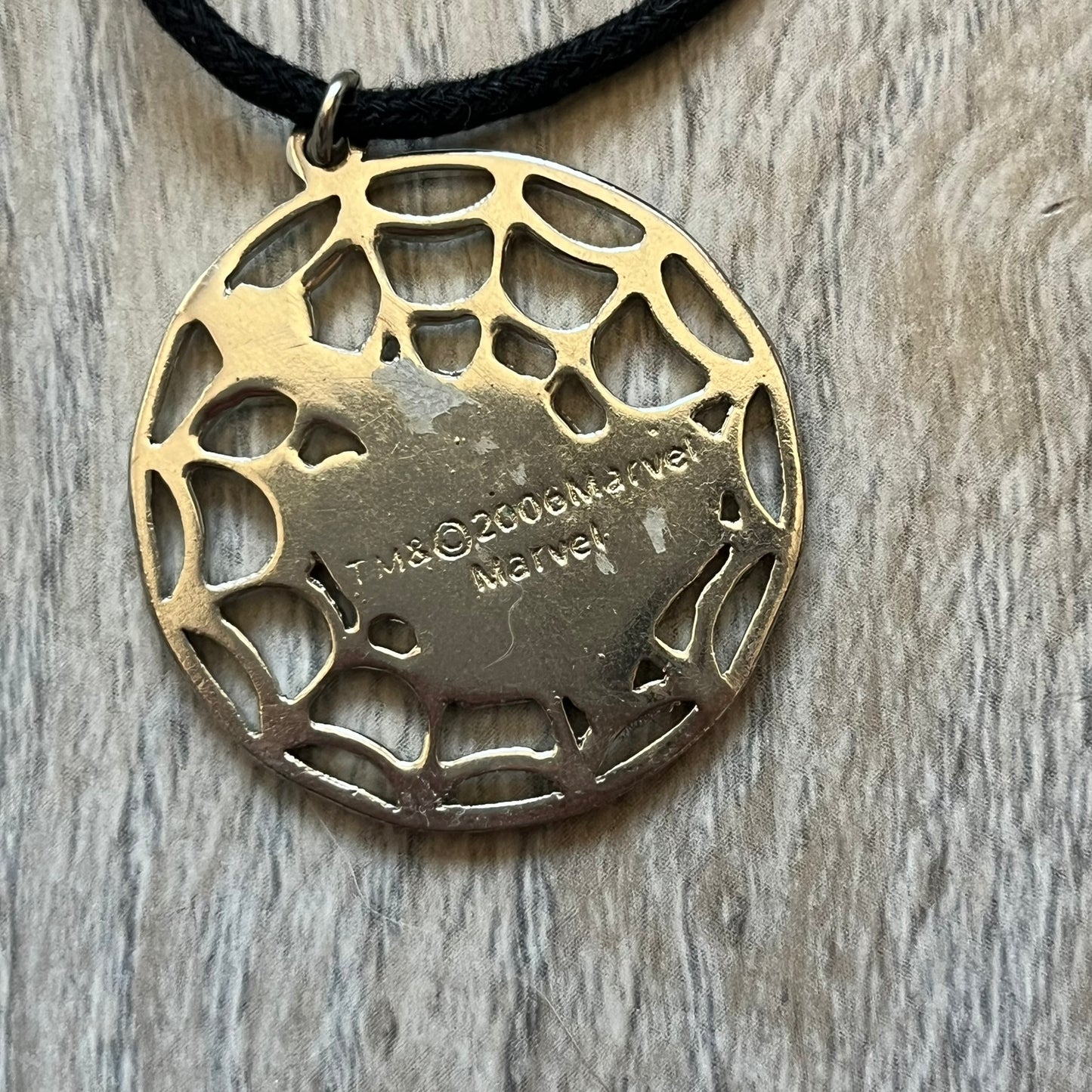 Spider-Man 2006 Web Silver Necklace is a necklace with a black cord and a circular silver-tone metal pendant. The front features a web background with a colored Spider-Man figure in a swinging pose, while the back is plain metal with 2006 licensing.