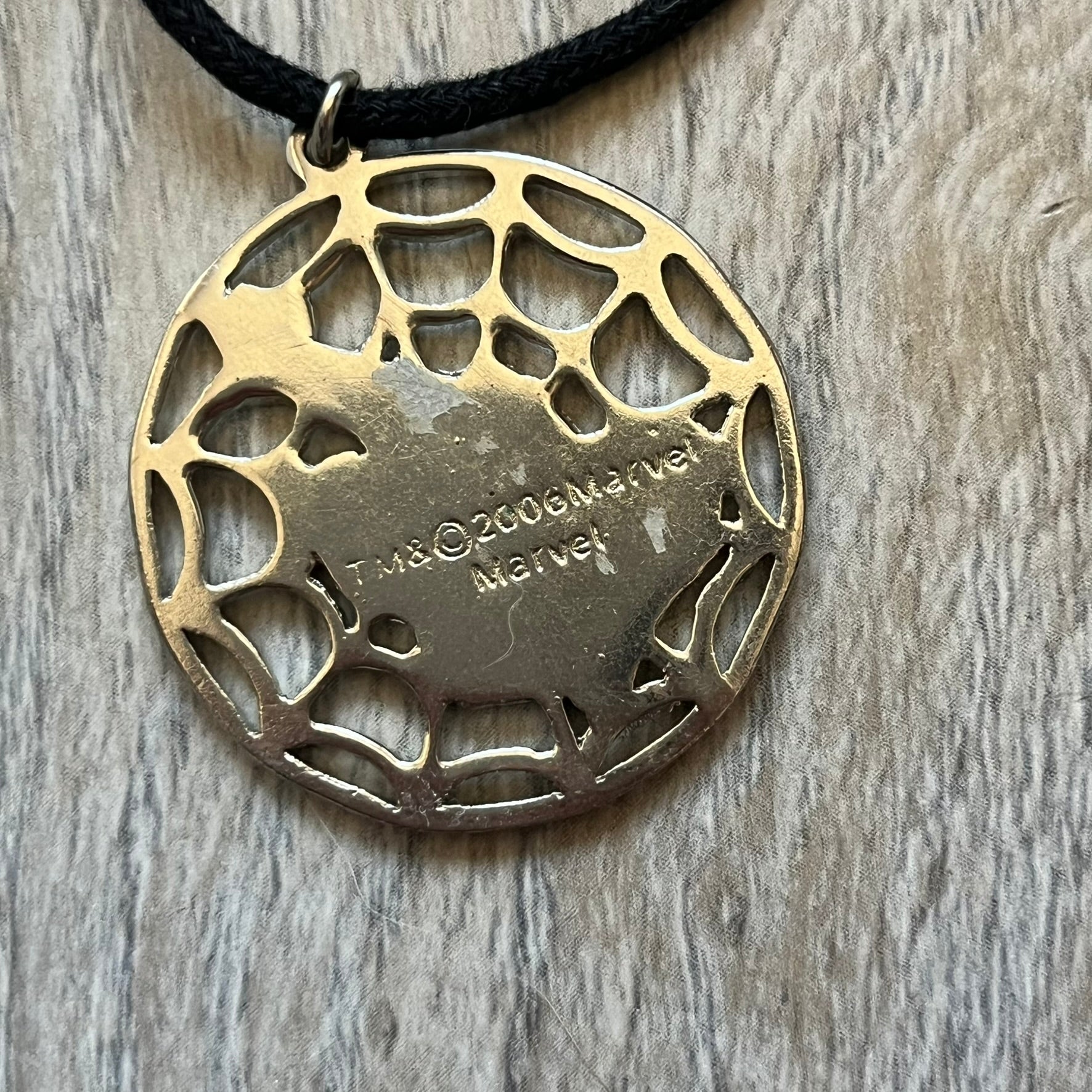 Spider-Man 2006 Web Silver Necklace is a necklace with a black cord and a circular silver-tone metal pendant. The front features a web background with a colored Spider-Man figure in a swinging pose, while the back is plain metal with 2006 licensing.