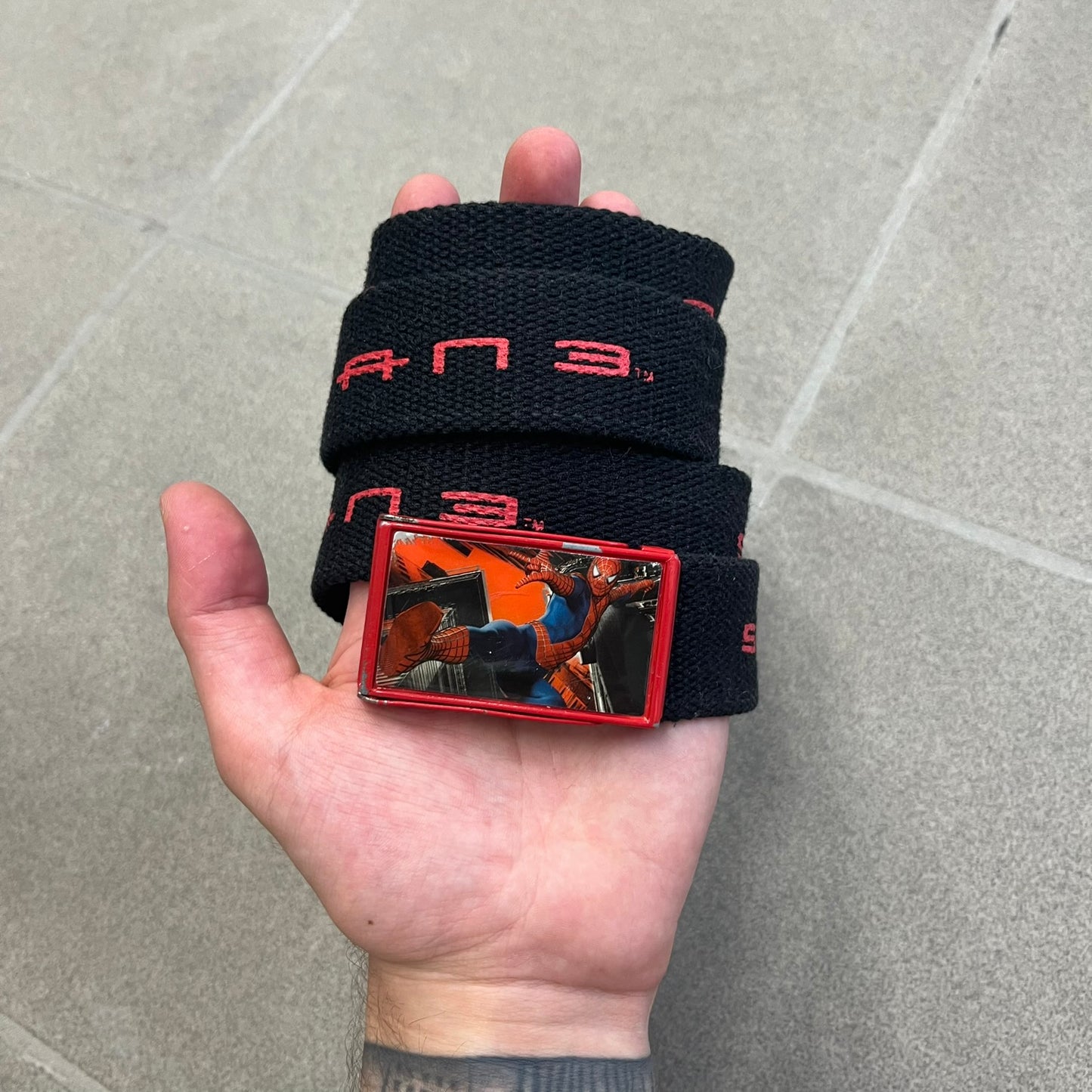 Spider-Man 3 2007 Black Belt is a black adjustable belt featuring repeated horizontal red Spider-Man 3 logo graphics across the strap. The belt has a red metal buckle with a Spider-Man graphic on the front showing him swinging. It includes a metal belt tip finish and a regulated sizing system for adjustment.
