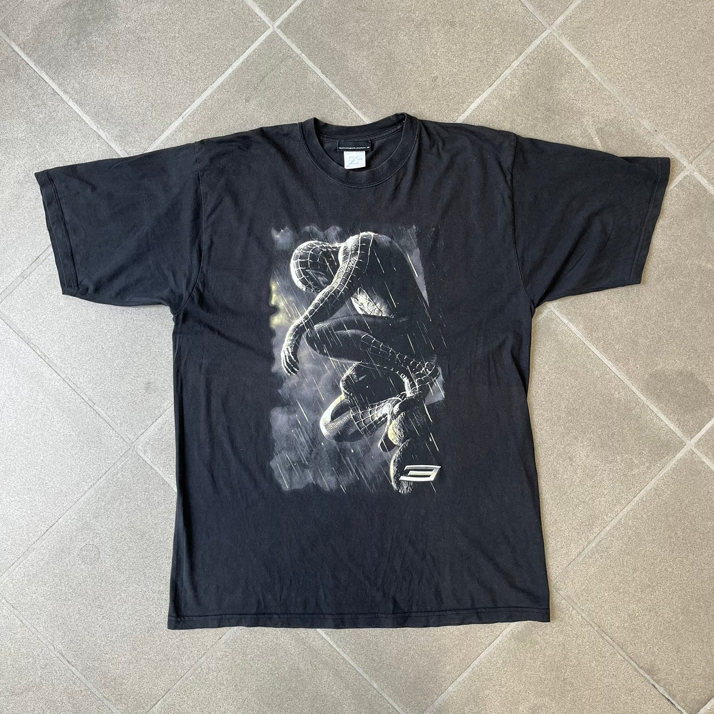 Spider-Man 3 Blacksuit 2007 Poster Tee features a black 100% cotton T-shirt with a large front graphic of Spider-Man crouching in the black suit in the rain. A “3” is printed in the bottom-right corner of the graphic. The shirt has a 2007 licensed black neck tag.

