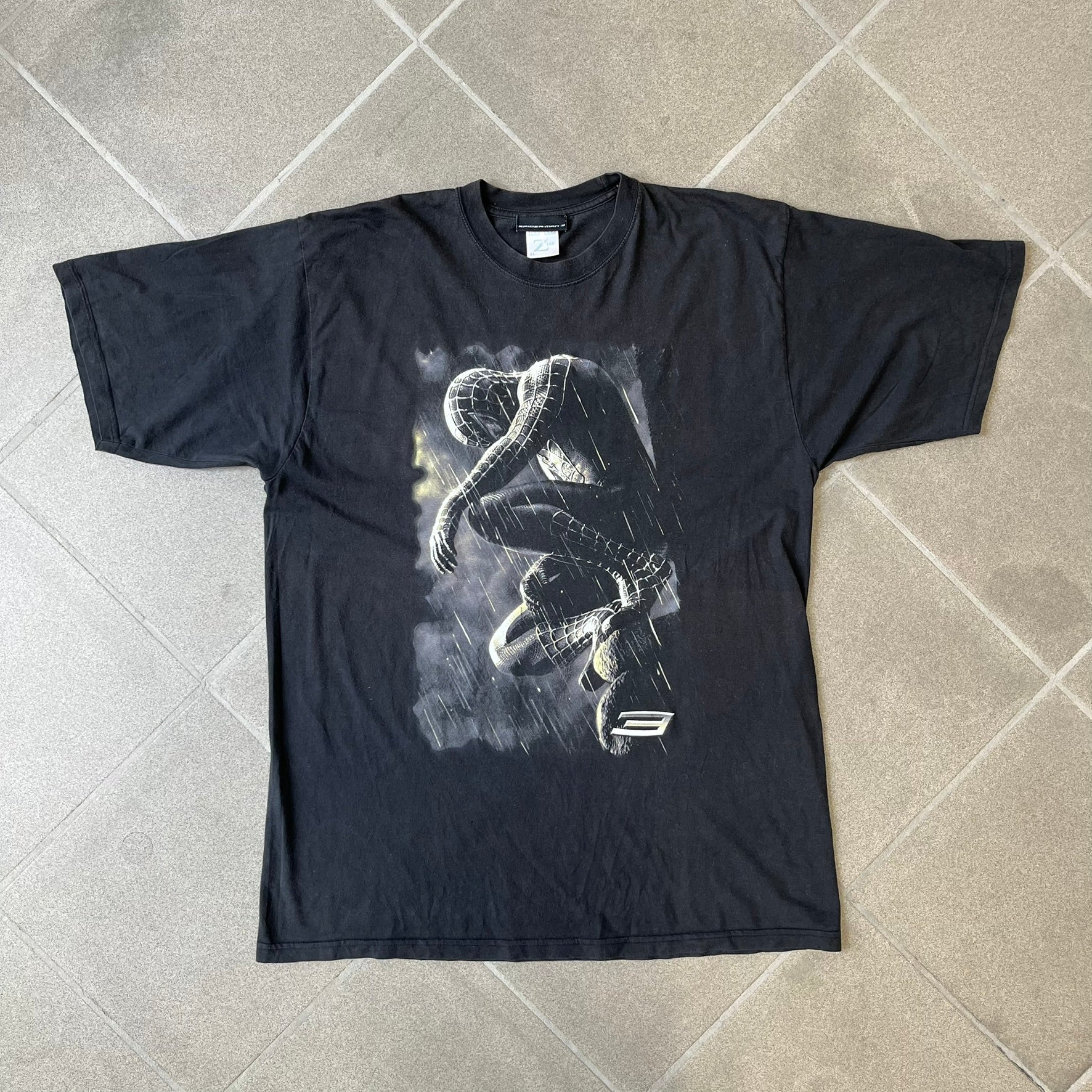 Spider-Man 3 Blacksuit 2007 Poster Tee features a black 100% cotton T-shirt with a large front graphic of Spider-Man crouching in the black suit in the rain. A “3” is printed in the bottom-right corner of the graphic. The shirt has a 2007 licensed black neck tag.
