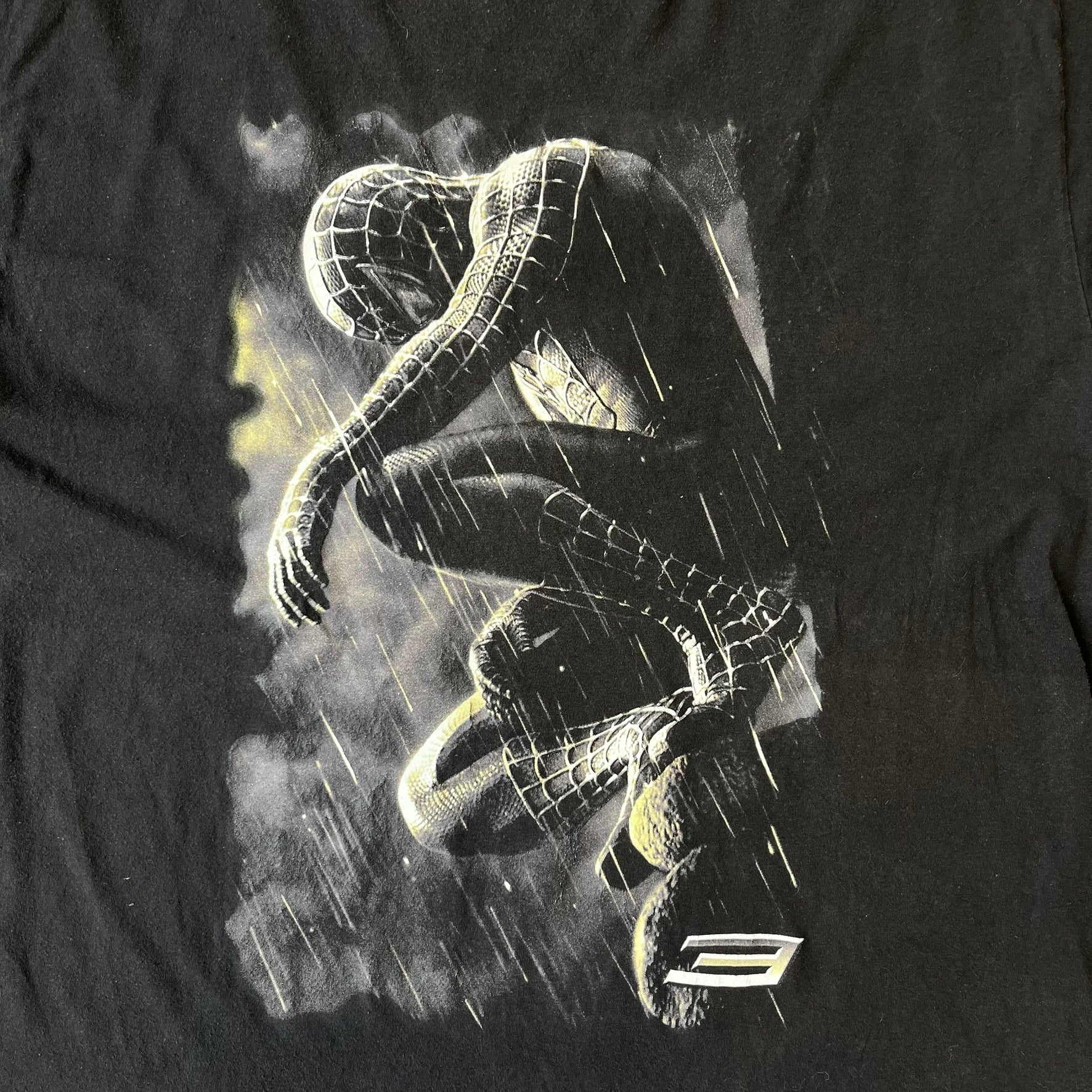 Spider-Man 3 Blacksuit 2007 Poster Tee features a black 100% cotton T-shirt with a large front graphic of Spider-Man crouching in the black suit in the rain. A “3” is printed in the bottom-right corner of the graphic. The shirt has a 2007 licensed black neck tag.
