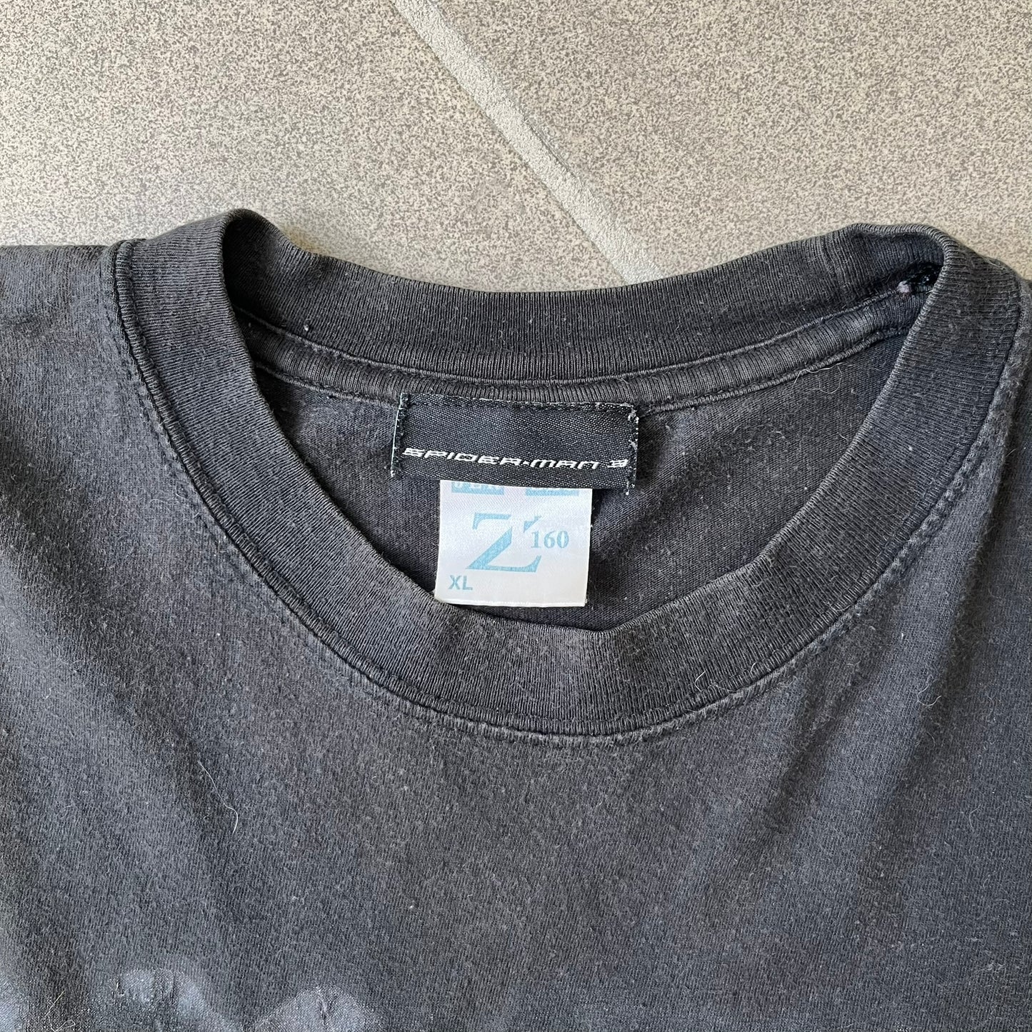 Spider-Man 3 Blacksuit 2007 Poster Tee features a black 100% cotton T-shirt with a large front graphic of Spider-Man crouching in the black suit in the rain. A “3” is printed in the bottom-right corner of the graphic. The shirt has a 2007 licensed black neck tag.
