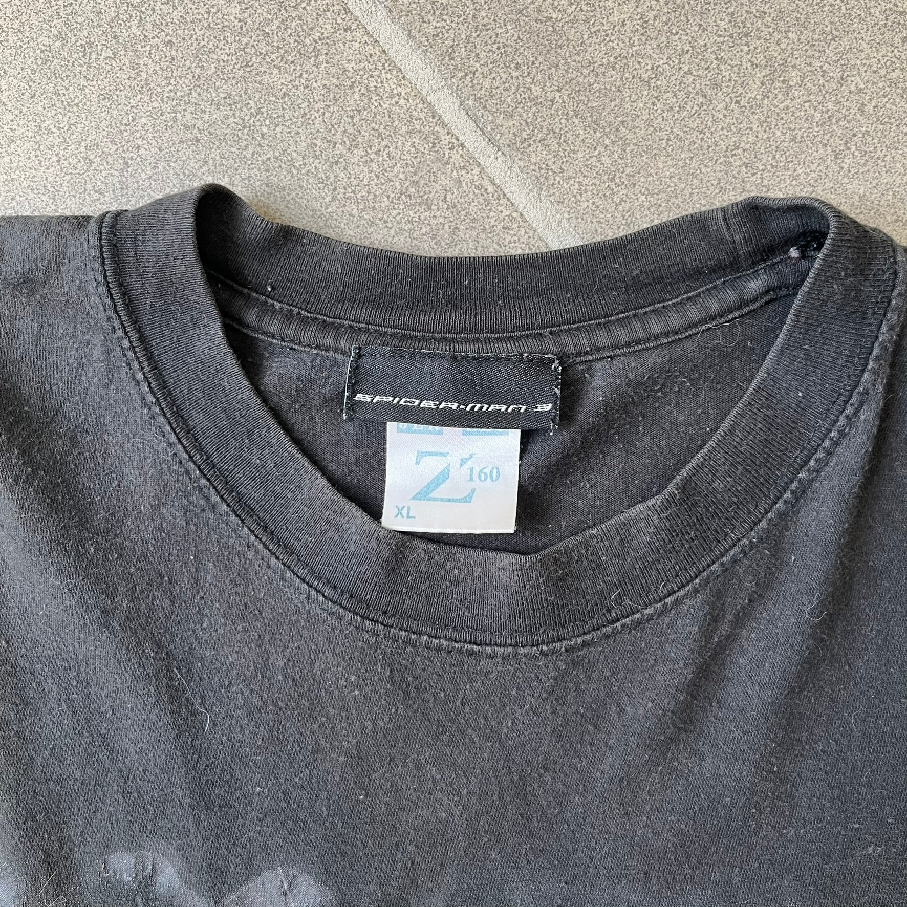Spider-Man 3 Blacksuit 2007 Poster Tee features a black 100% cotton T-shirt with a large front graphic of Spider-Man crouching in the black suit in the rain. A “3” is printed in the bottom-right corner of the graphic. The shirt has a 2007 licensed black neck tag.
