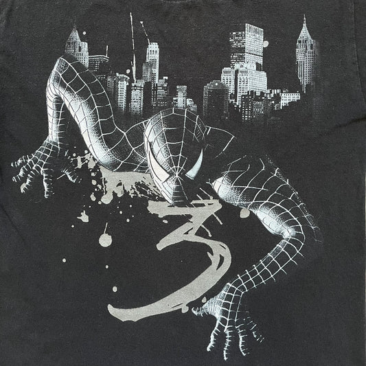 Spider-Man 3 Promo 2007 Black Tee is a black short-sleeve T-shirt with an original Spider-Man 3 neck tag. The front features a crawling black-suit Spider-Man graphic with a painted-style “3” beneath, set against a city background in white, grey, and black tones. The number “3” and the character’s eyes include a subtle glitter finish.
