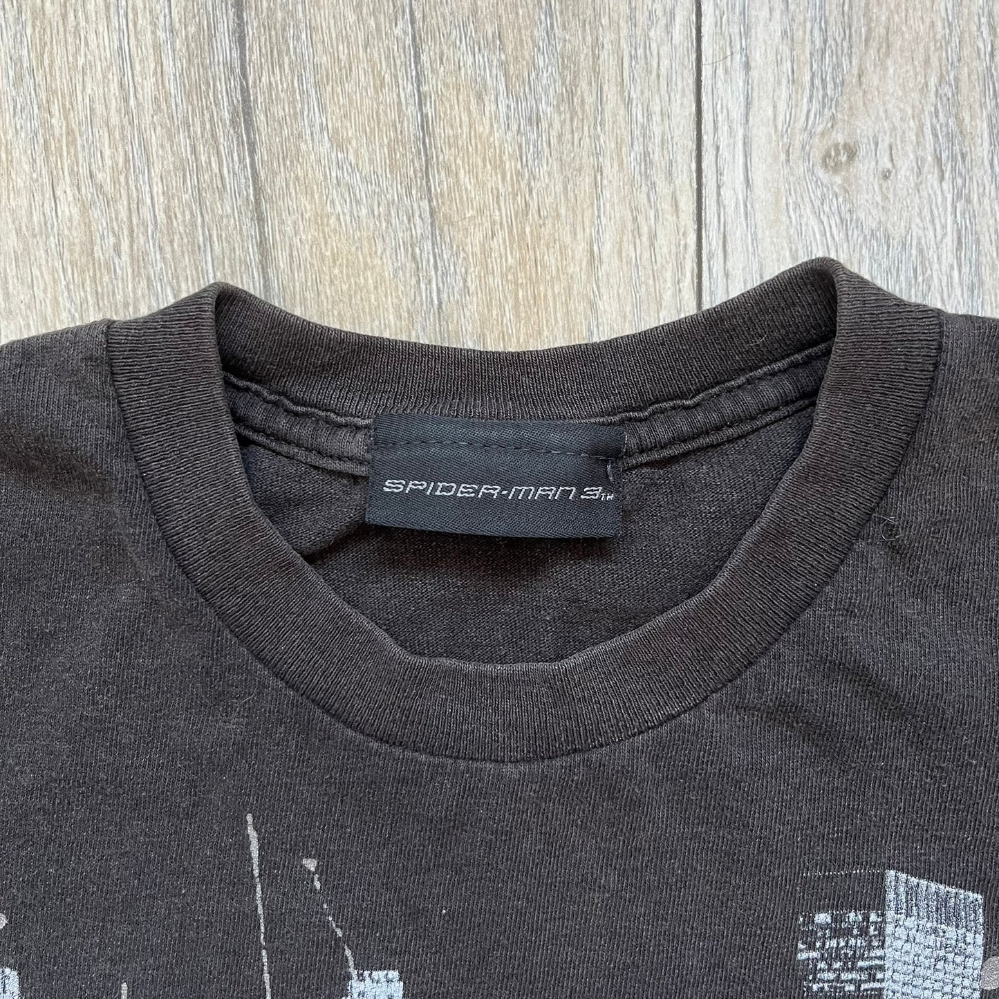 Spider-Man 3 Promo 2007 Black Tee is a black short-sleeve T-shirt with an original Spider-Man 3 neck tag. The front features a crawling black-suit Spider-Man graphic with a painted-style “3” beneath, set against a city background in white, grey, and black tones. The number “3” and the character’s eyes include a subtle glitter finish.
