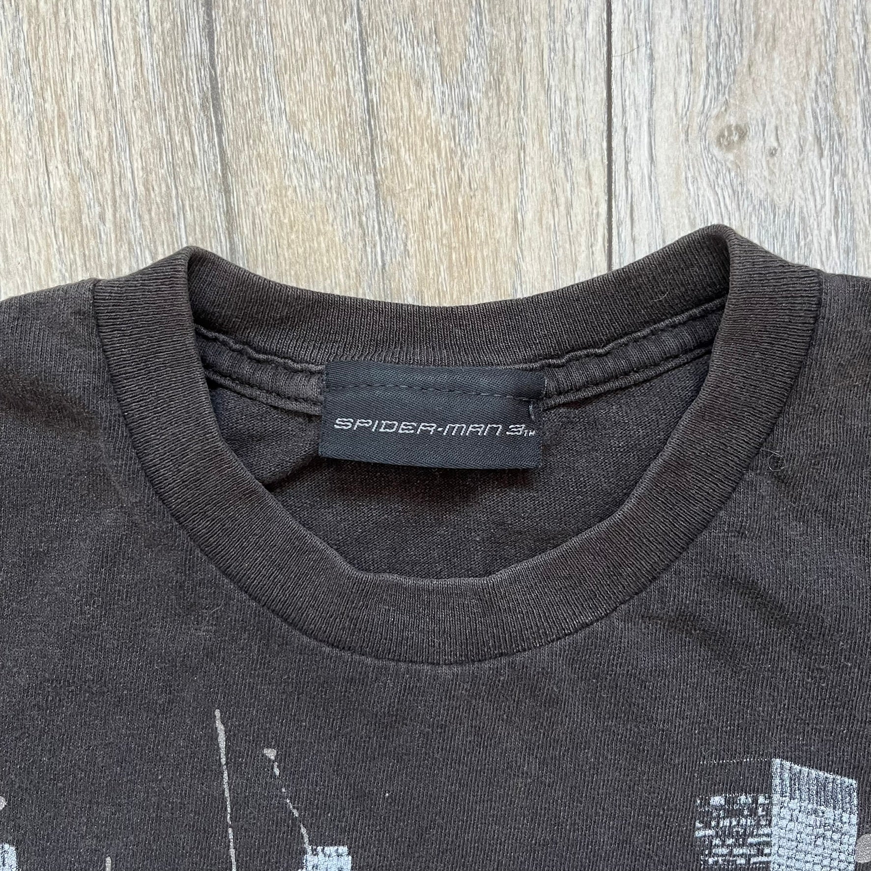 Spider-Man 3 Promo 2007 Black Tee is a black short-sleeve T-shirt with an original Spider-Man 3 neck tag. The front features a crawling black-suit Spider-Man graphic with a painted-style “3” beneath, set against a city background in white, grey, and black tones. The number “3” and the character’s eyes include a subtle glitter finish.
