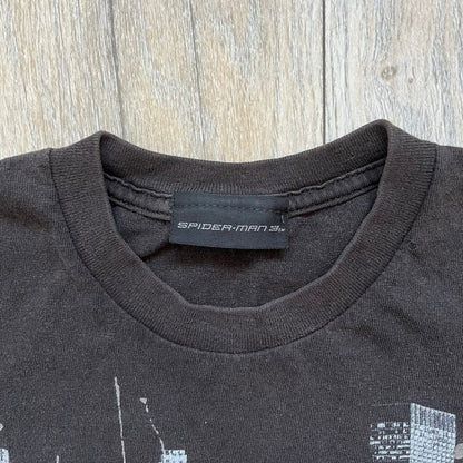 Spider-Man 3 Promo 2007 Black Tee is a black short-sleeve T-shirt with an original Spider-Man 3 neck tag. The front features a crawling black-suit Spider-Man graphic with a painted-style “3” beneath, set against a city background in white, grey, and black tones. The number “3” and the character’s eyes include a subtle glitter finish.
