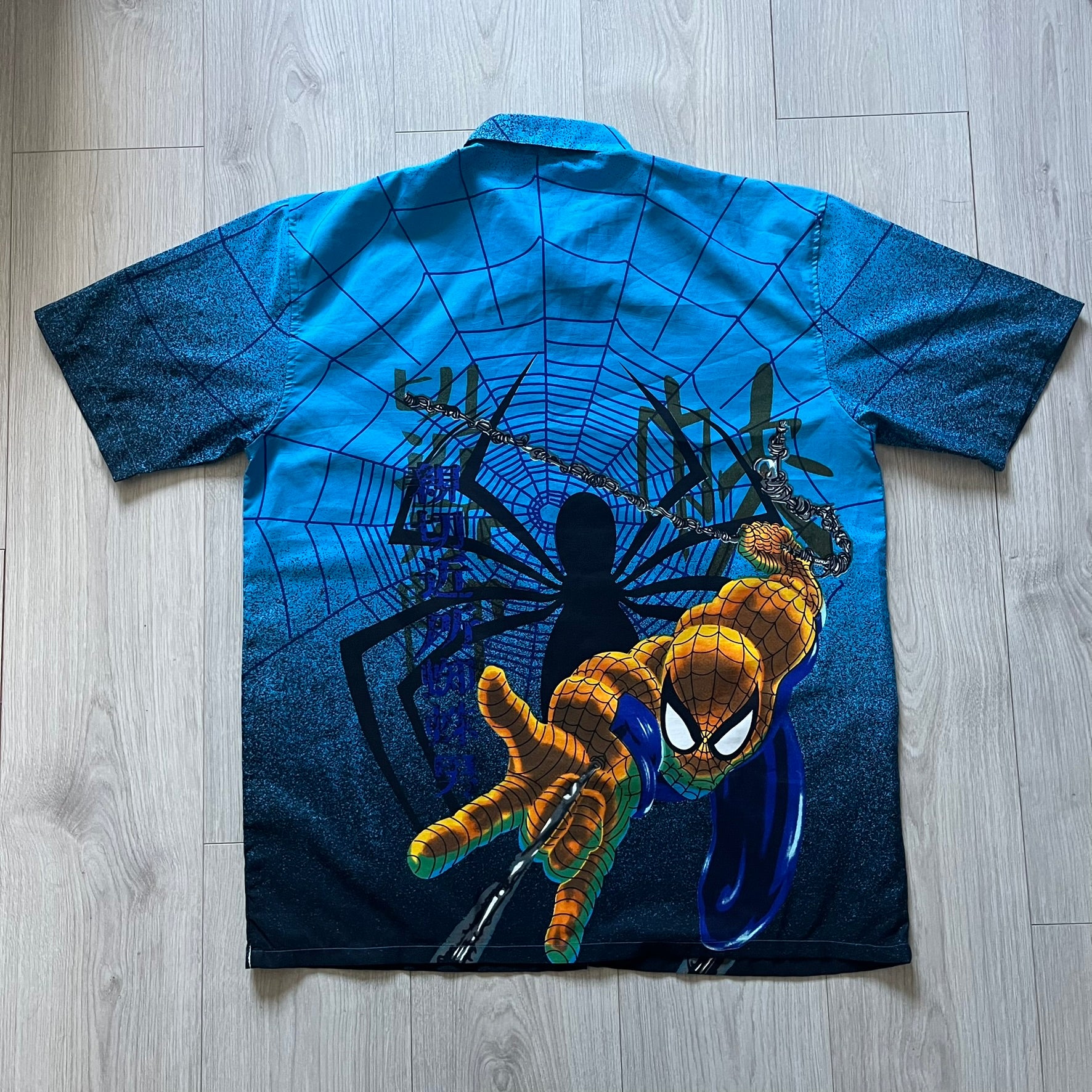 Blue Spider-Man button-up shirt featuring an all-over web print and mirrored graphics of Spider-Man shooting webs on both sides. Centered Spider-Man logo completes the great design. A striking, comic-inspired piece perfect for fans and collectors.