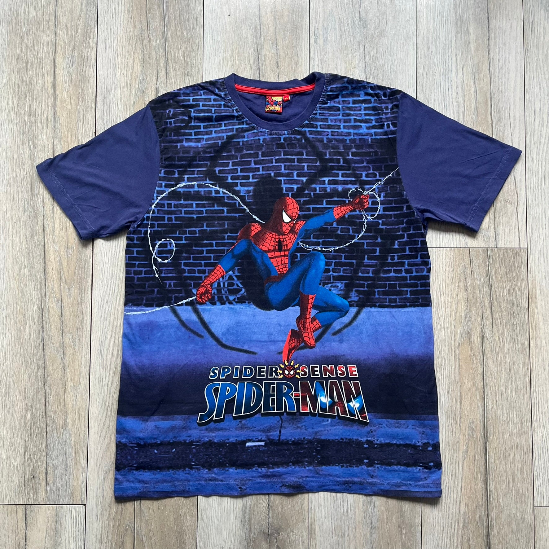 Spider-Man “Spider Sense” 2010 AOP Tee is a navy all-over print style T-shirt featuring a large front graphic of cartoon Spider-Man swinging in front of a brick wall with a black Spider-Man logo mural. Below the artwork is detailed “Spider Sense Spider-Man” text. The back features a simple outlined white Spider-Man logo near the upper back. Produced under license by Contrast with 2010 Marvel licensing and a detailed branded neck tag.
