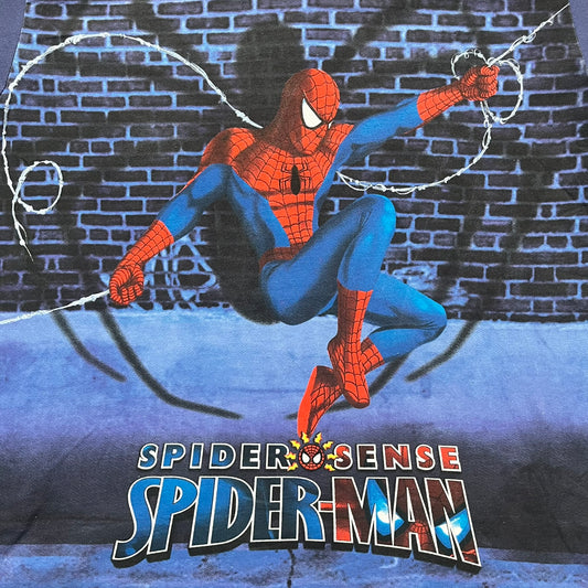 Spider-Man “Spider Sense” 2010 AOP Tee is a navy all-over print style T-shirt featuring a large front graphic of cartoon Spider-Man swinging in front of a brick wall with a black Spider-Man logo mural. Below the artwork is detailed “Spider Sense Spider-Man” text. The back features a simple outlined white Spider-Man logo near the upper back. Produced under license by Contrast with 2010 Marvel licensing and a detailed branded neck tag.
