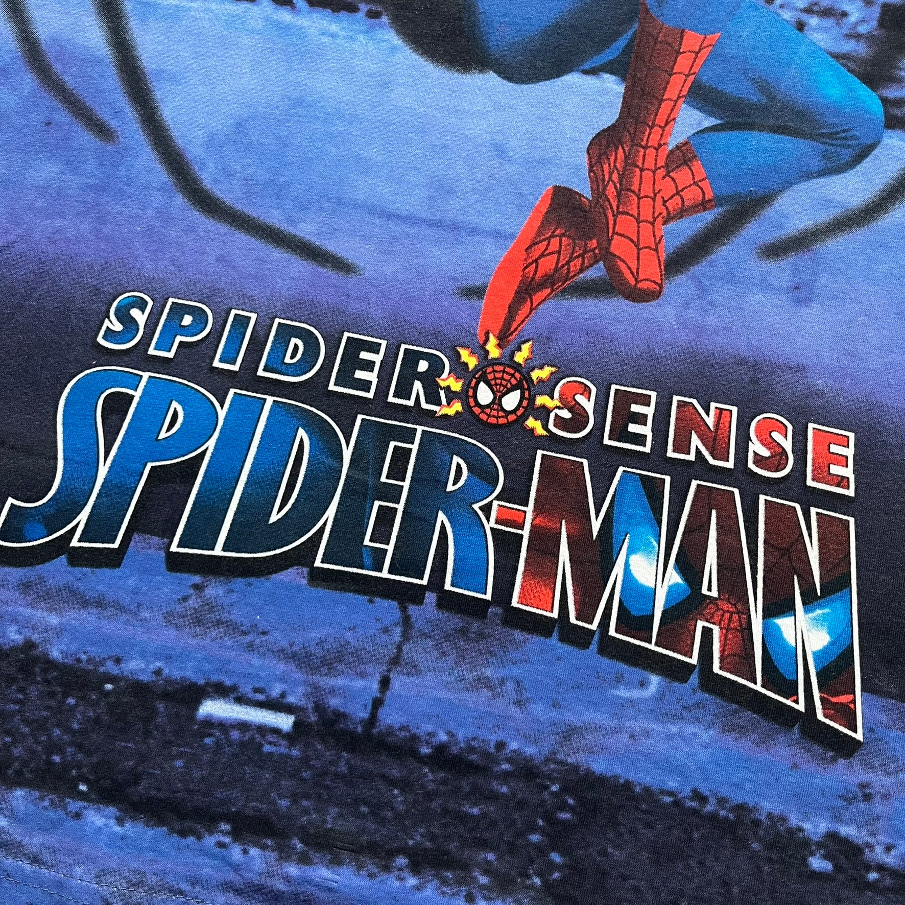Spider-Man “Spider Sense” 2010 AOP Tee is a navy all-over print style T-shirt featuring a large front graphic of cartoon Spider-Man swinging in front of a brick wall with a black Spider-Man logo mural. Below the artwork is detailed “Spider Sense Spider-Man” text. The back features a simple outlined white Spider-Man logo near the upper back. Produced under license by Contrast with 2010 Marvel licensing and a detailed branded neck tag.
