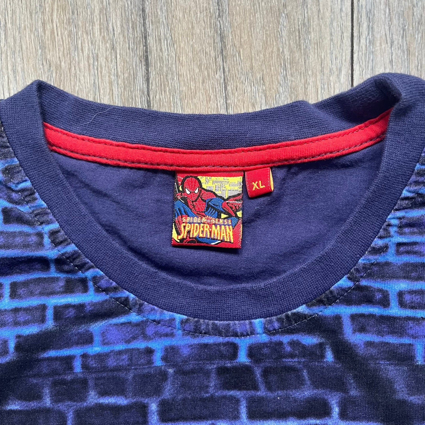 Spider-Man “Spider Sense” 2010 AOP Tee is a navy all-over print style T-shirt featuring a large front graphic of cartoon Spider-Man swinging in front of a brick wall with a black Spider-Man logo mural. Below the artwork is detailed “Spider Sense Spider-Man” text. The back features a simple outlined white Spider-Man logo near the upper back. Produced under license by Contrast with 2010 Marvel licensing and a detailed branded neck tag.
