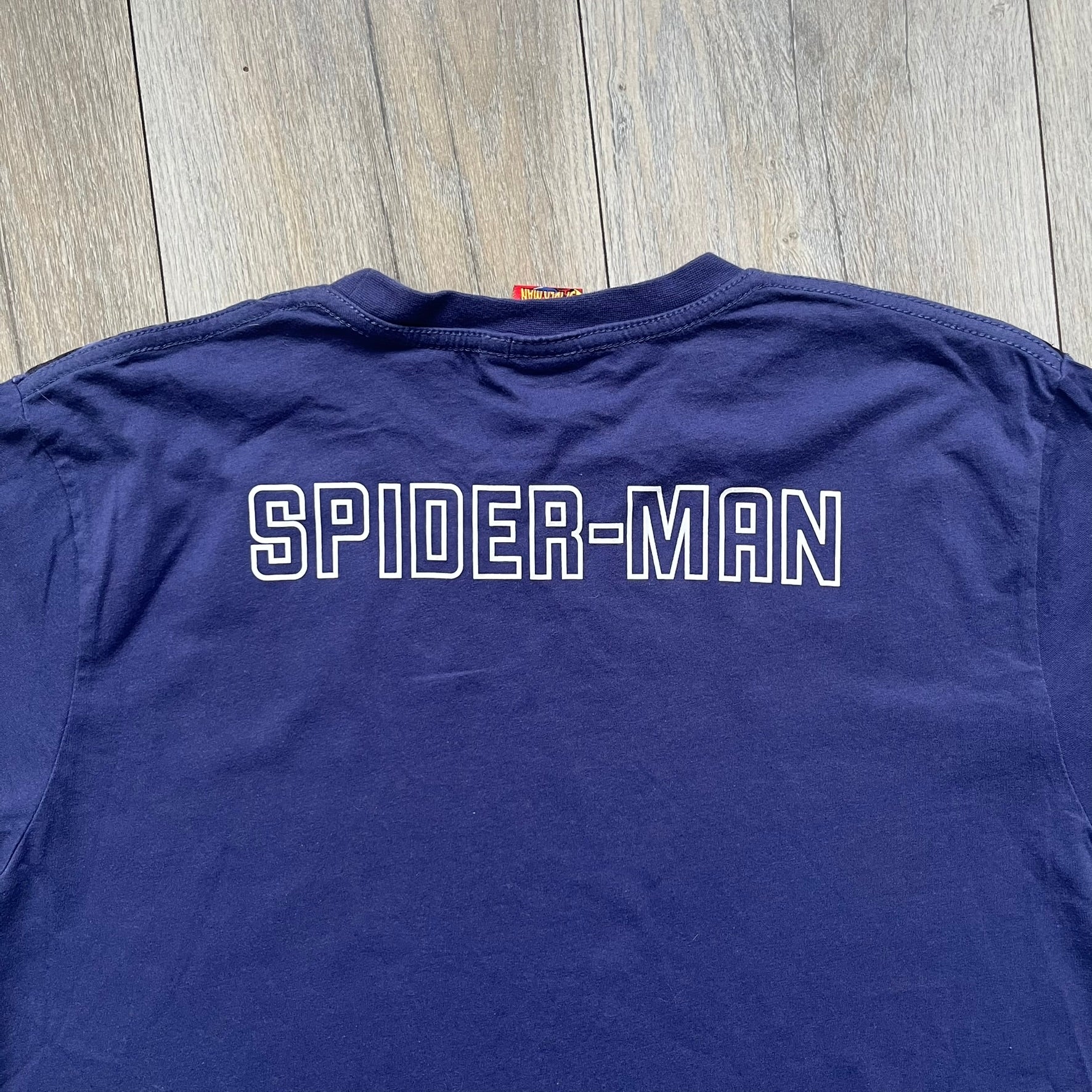 Spider-Man “Spider Sense” 2010 AOP Tee is a navy all-over print style T-shirt featuring a large front graphic of cartoon Spider-Man swinging in front of a brick wall with a black Spider-Man logo mural. Below the artwork is detailed “Spider Sense Spider-Man” text. The back features a simple outlined white Spider-Man logo near the upper back. Produced under license by Contrast with 2010 Marvel licensing and a detailed branded neck tag.
