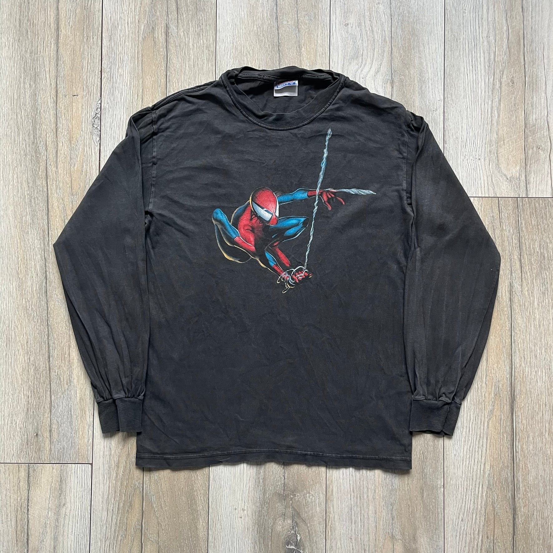 Spider-Man Swinging 2003 Washed Longsleeve is a washed black and dark gray cotton long-sleeve shirt made on a Hanes Beefy-T blank. The front features a large detailed comic-style Spider-Man graphic in red and blue, showing the character swinging in motion. Made from 100% cotton with a faded vintage finish.
