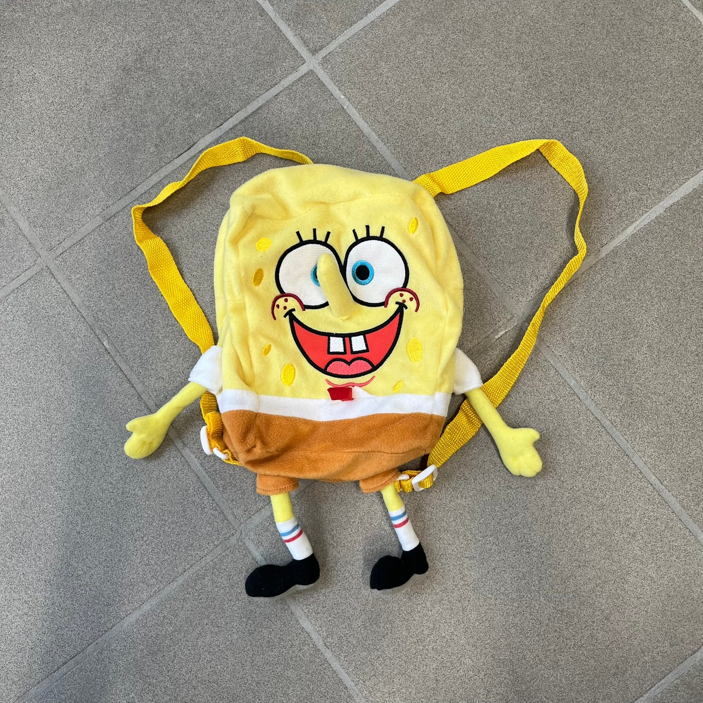 SpongeBob SquarePants 2010 Backpack is designed in the shape of SpongeBob’s body with embroidered facial details, a 3D nose, and loose hanging arms and legs. It features one main compartment and a yellow 2010 licensed tag on the side.
