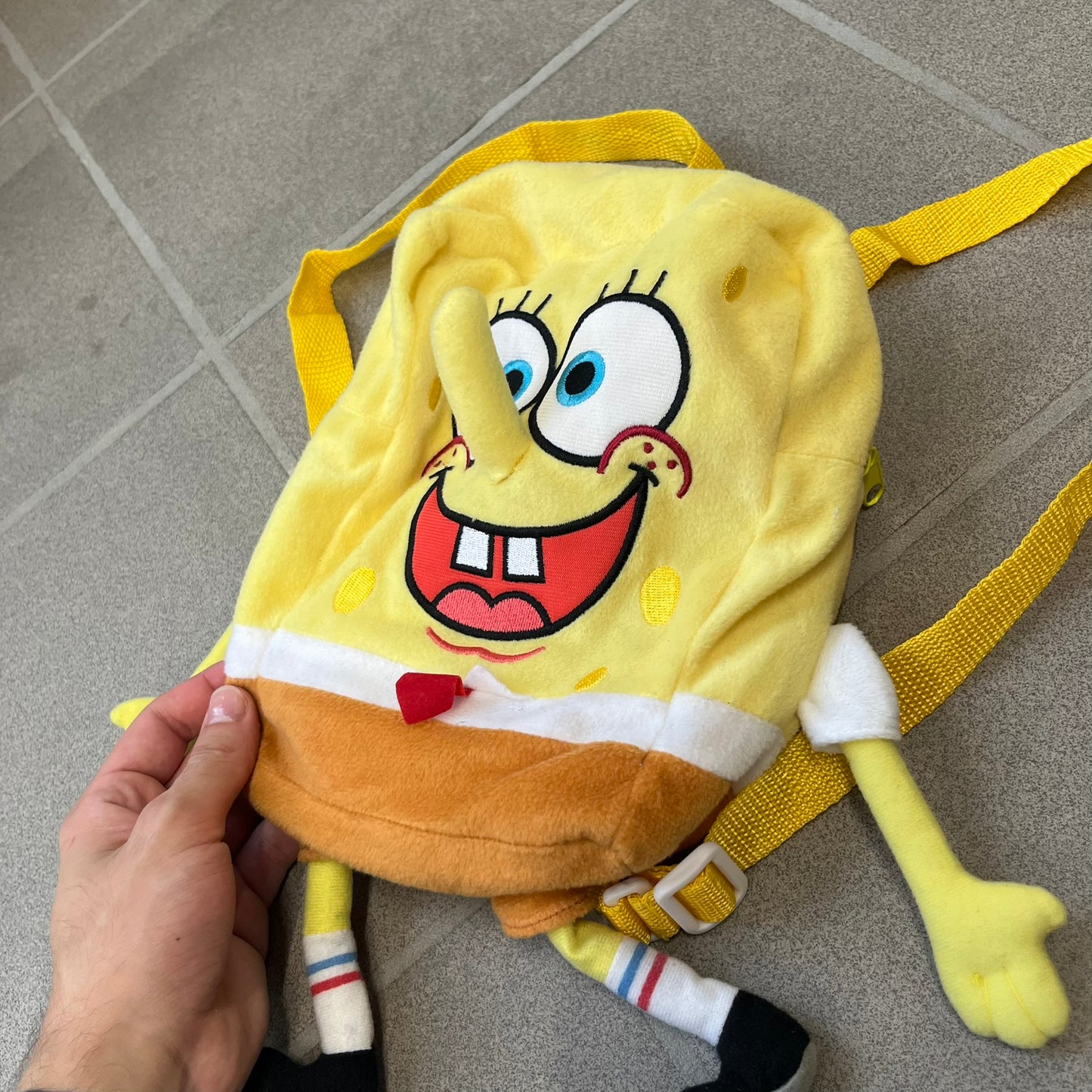 SpongeBob SquarePants 2010 Backpack is designed in the shape of SpongeBob’s body with embroidered facial details, a 3D nose, and loose hanging arms and legs. It features one main compartment and a yellow 2010 licensed tag on the side.
