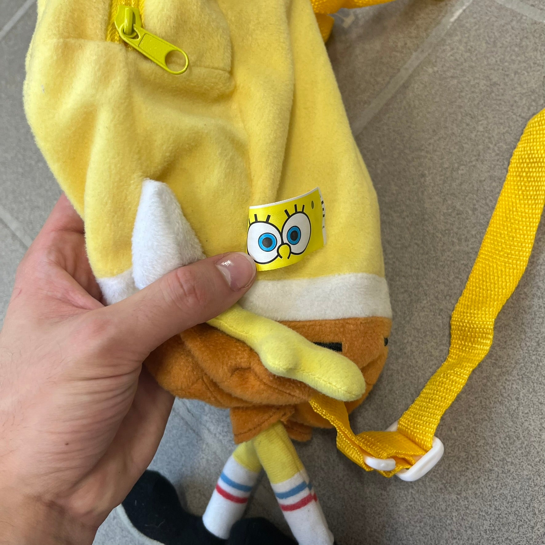 SpongeBob SquarePants 2010 Backpack is designed in the shape of SpongeBob’s body with embroidered facial details, a 3D nose, and loose hanging arms and legs. It features one main compartment and a yellow 2010 licensed tag on the side.
