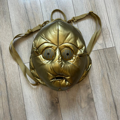 Star Wars 1990s C-3PO Head Backpack is a novelty backpack shaped like the head of C-3PO. It features a metallic gold finish with detailed sculpted paneling and plastic lens-style eyes for a robotic look. The bag includes a 1990s Star Wars red licensed tag and a single main compartment.