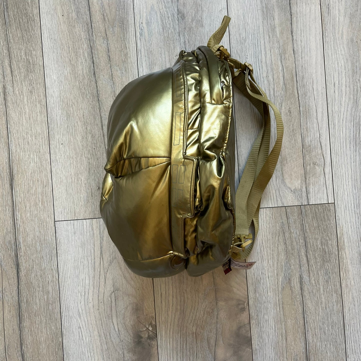 Star Wars 1990s C-3PO Head Backpack is a novelty backpack shaped like the head of C-3PO. It features a metallic gold finish with detailed sculpted paneling and plastic lens-style eyes for a robotic look. The bag includes a 1990s Star Wars red licensed tag and a single main compartment.
