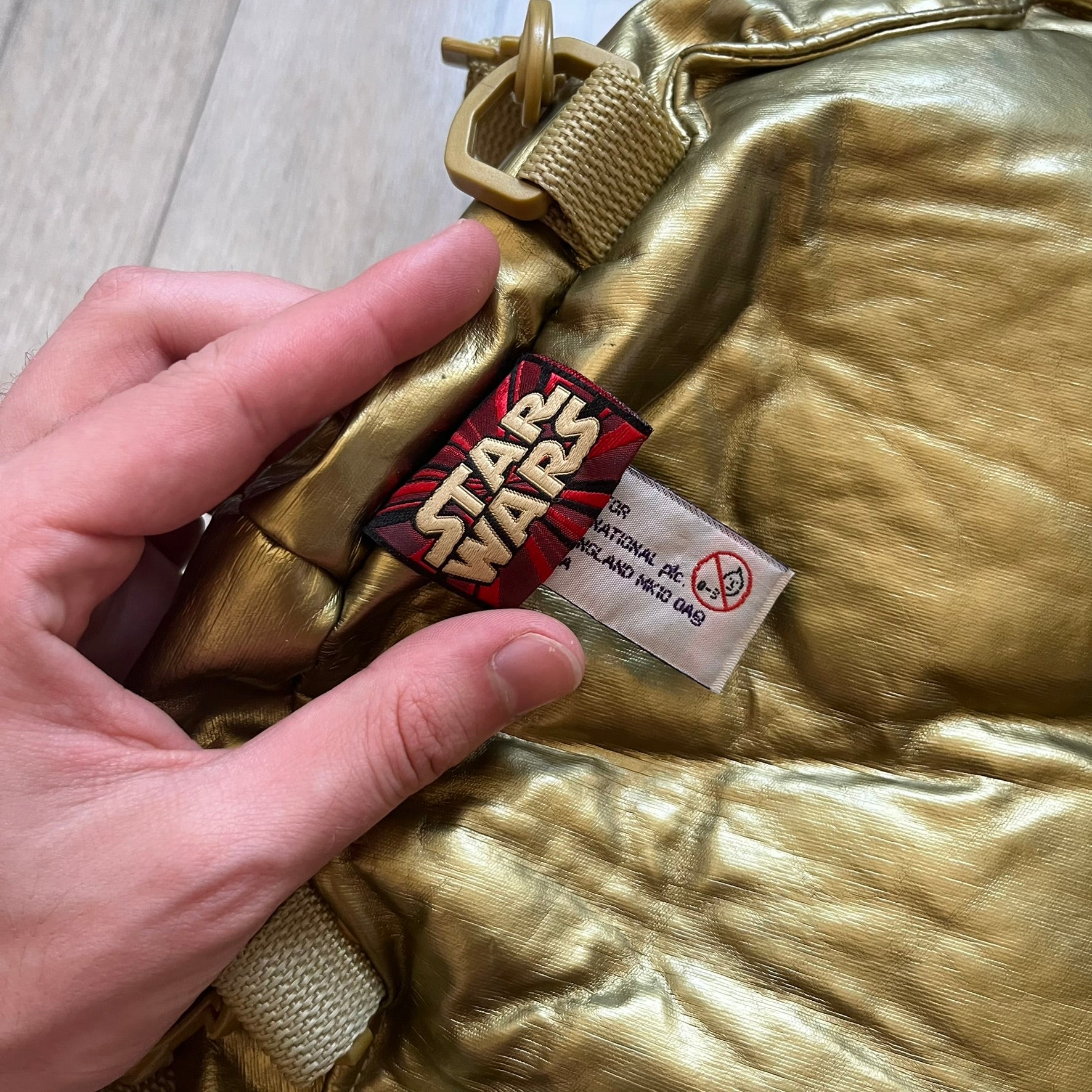Star Wars 1990s C-3PO Head Backpack is a novelty backpack shaped like the head of C-3PO. It features a metallic gold finish with detailed sculpted paneling and plastic lens-style eyes for a robotic look. The bag includes a 1990s Star Wars red licensed tag and a single main compartment.