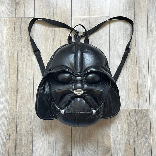 **Star Wars 1990s Darth Vader Head Backpack** is a vintage backpack shaped like Darth Vader’s helmet. It is finished in shiny black with molded plastic details recreating the character’s mask and helmet. The bag has one main compartment and includes a red Star Wars licensed tag from the 1990s.
