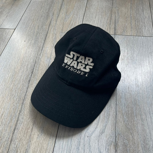 Star Wars Episode I 1990s Black Baseball Cap is a black cotton cap featuring embroidered Star Wars branding on the front in white and gray tones. Beneath it, a smaller “Episode I” embroidery is included for additional detailing. The interior includes a red Star Wars licensed tag, completing the official branded construction.