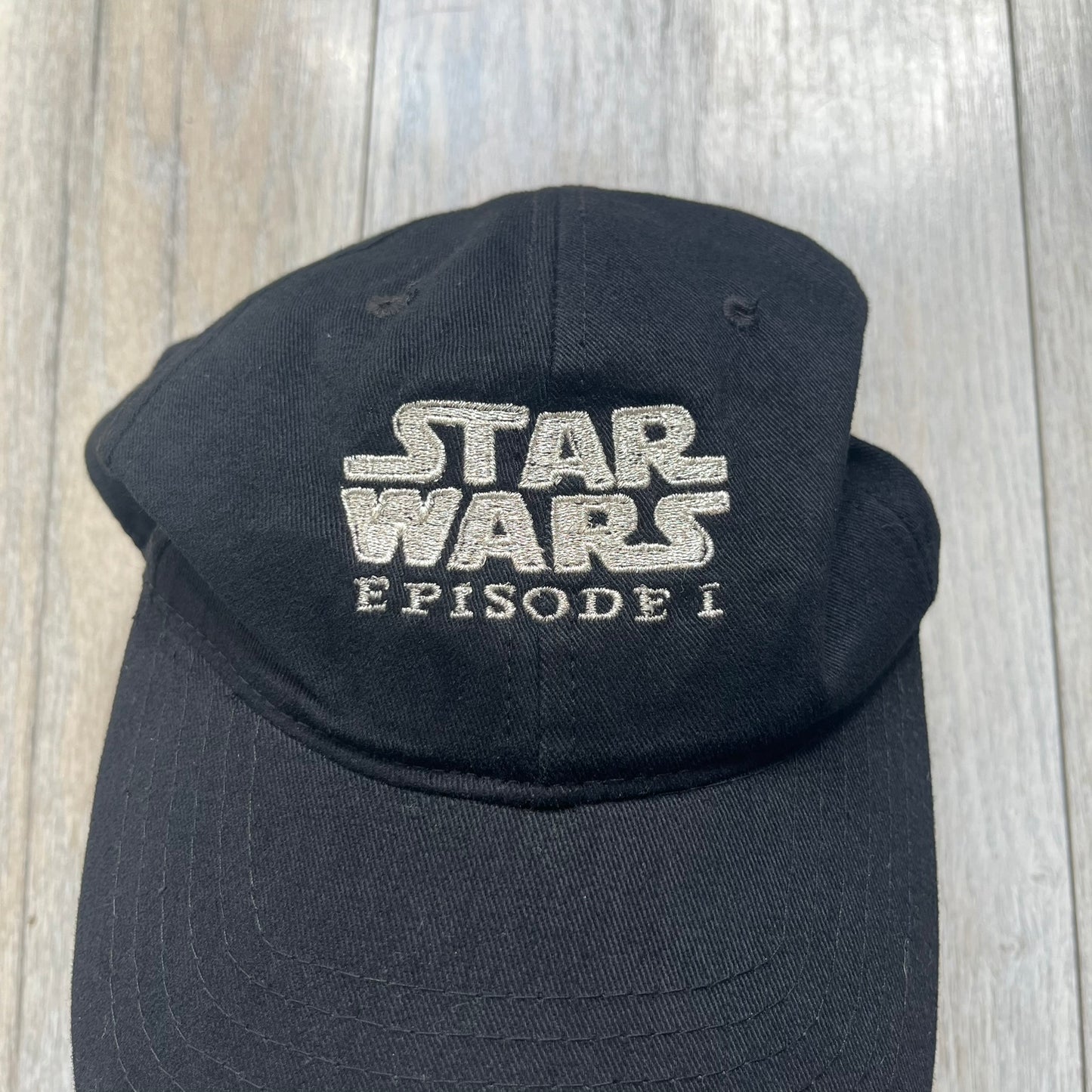 Star Wars Episode I 1990s Black Baseball Cap is a black cotton cap featuring embroidered Star Wars branding on the front in white and gray tones. Beneath it, a smaller “Episode I” embroidery is included for additional detailing. The interior includes a red Star Wars licensed tag, completing the official branded construction.