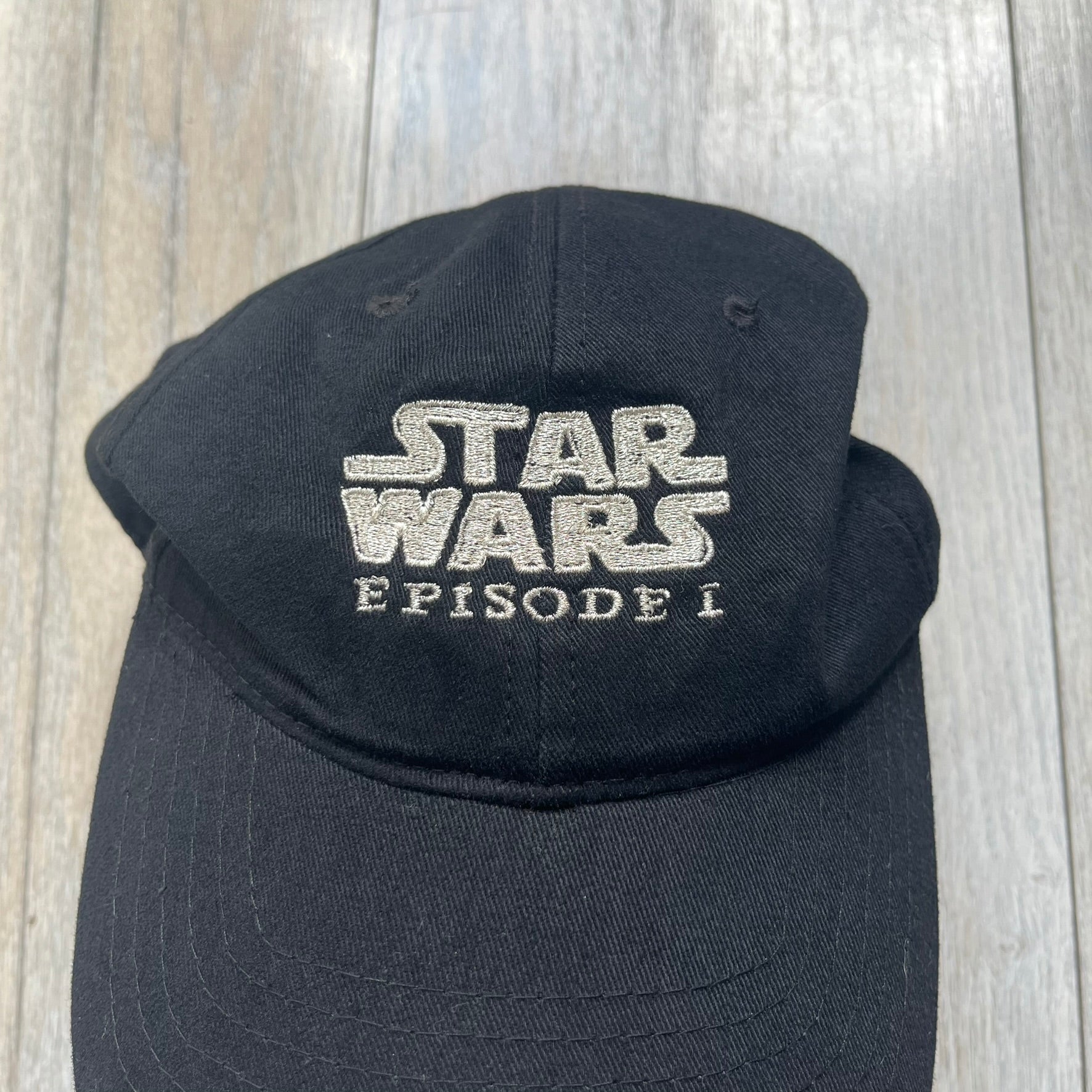 Star Wars Episode I 1990s Black Baseball Cap is a black cotton cap featuring embroidered Star Wars branding on the front in white and gray tones. Beneath it, a smaller “Episode I” embroidery is included for additional detailing. The interior includes a red Star Wars licensed tag, completing the official branded construction.
