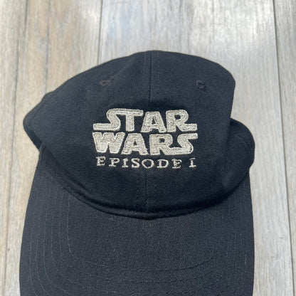 Star Wars Episode I 1990s Black Baseball Cap is a black cotton cap featuring embroidered Star Wars branding on the front in white and gray tones. Beneath it, a smaller “Episode I” embroidery is included for additional detailing. The interior includes a red Star Wars licensed tag, completing the official branded construction.