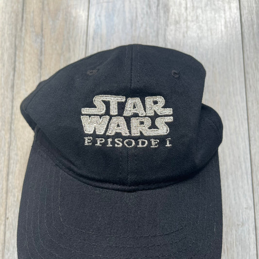 Star Wars Episode I 1990s Black Baseball Cap is a black cotton cap featuring embroidered Star Wars branding on the front in white and gray tones. Beneath it, a smaller “Episode I” embroidery is included for additional detailing. The interior includes a red Star Wars licensed tag, completing the official branded construction.