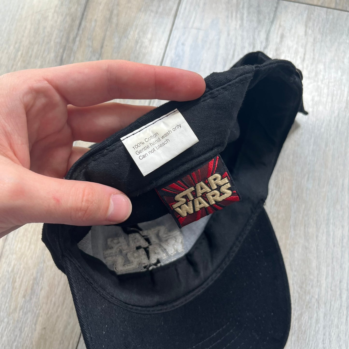 Star Wars Episode I 1990s Black Baseball Cap is a black cotton cap featuring embroidered Star Wars branding on the front in white and gray tones. Beneath it, a smaller “Episode I” embroidery is included for additional detailing. The interior includes a red Star Wars licensed tag, completing the official branded construction.