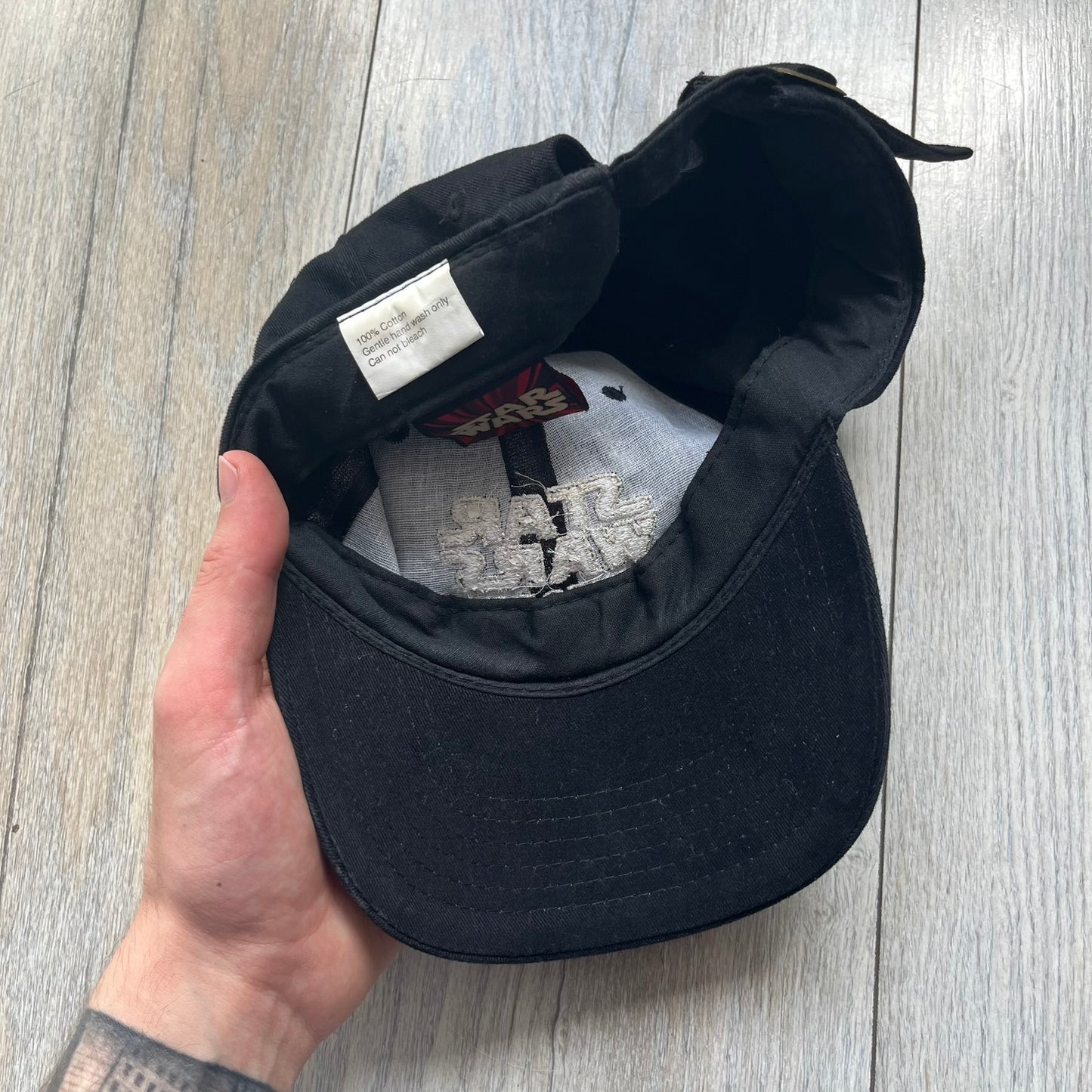 Star Wars Episode I 1990s Black Baseball Cap is a black cotton cap featuring embroidered Star Wars branding on the front in white and gray tones. Beneath it, a smaller “Episode I” embroidery is included for additional detailing. The interior includes a red Star Wars licensed tag, completing the official branded construction.