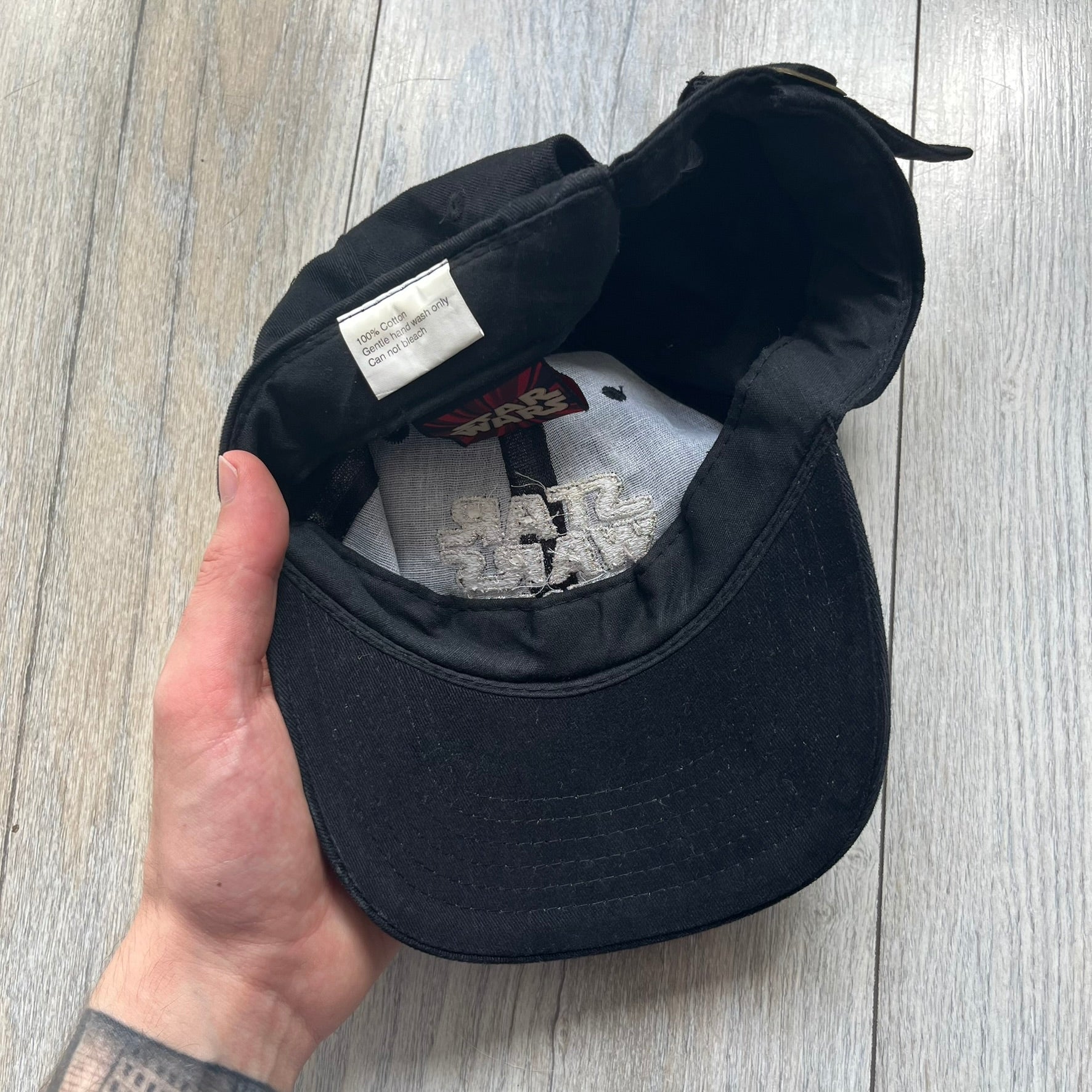 Star Wars Episode I 1990s Black Baseball Cap is a black cotton cap featuring embroidered Star Wars branding on the front in white and gray tones. Beneath it, a smaller “Episode I” embroidery is included for additional detailing. The interior includes a red Star Wars licensed tag, completing the official branded construction.