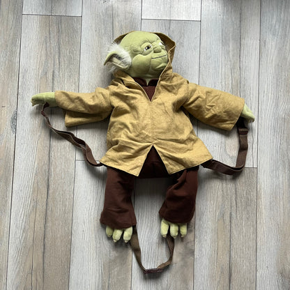 Star Wars Episode III Yoda 2006 Backpack is a character-shaped backpack featuring Yoda dressed in brown pants and a beige-yellow jacket with an attached hood. It includes two standard shoulder straps running from the hands to the feet, plus an additional stabilizing strap attached near the legs. The interior tag shows Star Wars Episode III: Revenge of the Sith branding with 2006 Lucasfilm licensing.
