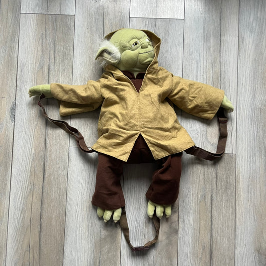 Star Wars Episode III Yoda 2006 Backpack is a character-shaped backpack featuring Yoda dressed in brown pants and a beige-yellow jacket with an attached hood. It includes two standard shoulder straps running from the hands to the feet, plus an additional stabilizing strap attached near the legs. The interior tag shows Star Wars Episode III: Revenge of the Sith branding with 2006 Lucasfilm licensing.