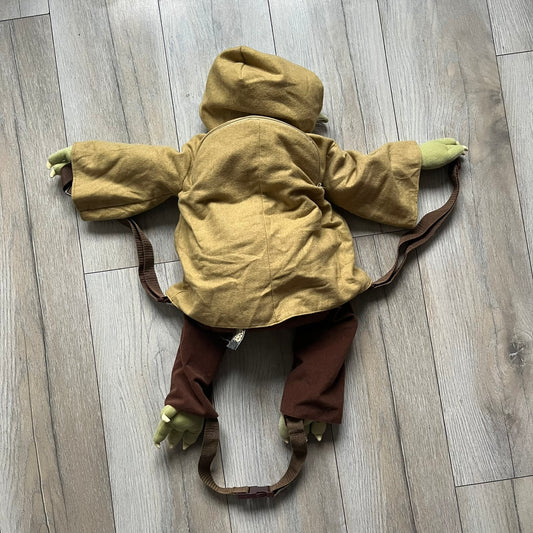 Star Wars Episode III Yoda 2006 Backpack is a character-shaped backpack featuring Yoda dressed in brown pants and a beige-yellow jacket with an attached hood. It includes two standard shoulder straps running from the hands to the feet, plus an additional stabilizing strap attached near the legs. The interior tag shows Star Wars Episode III: Revenge of the Sith branding with 2006 Lucasfilm licensing.