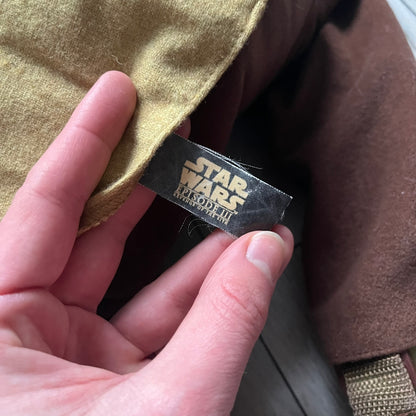 Star Wars Episode III Yoda 2006 Backpack is a character-shaped backpack featuring Yoda dressed in brown pants and a beige-yellow jacket with an attached hood. It includes two standard shoulder straps running from the hands to the feet, plus an additional stabilizing strap attached near the legs. The interior tag shows Star Wars Episode III: Revenge of the Sith branding with 2006 Lucasfilm licensing.