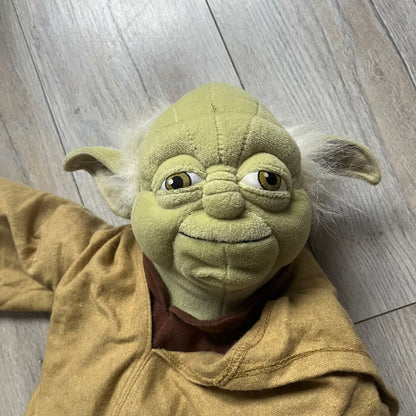 Star Wars Episode III Yoda 2006 Backpack is a character-shaped backpack featuring Yoda dressed in brown pants and a beige-yellow jacket with an attached hood. It includes two standard shoulder straps running from the hands to the feet, plus an additional stabilizing strap attached near the legs. The interior tag shows Star Wars Episode III: Revenge of the Sith branding with 2006 Lucasfilm licensing.