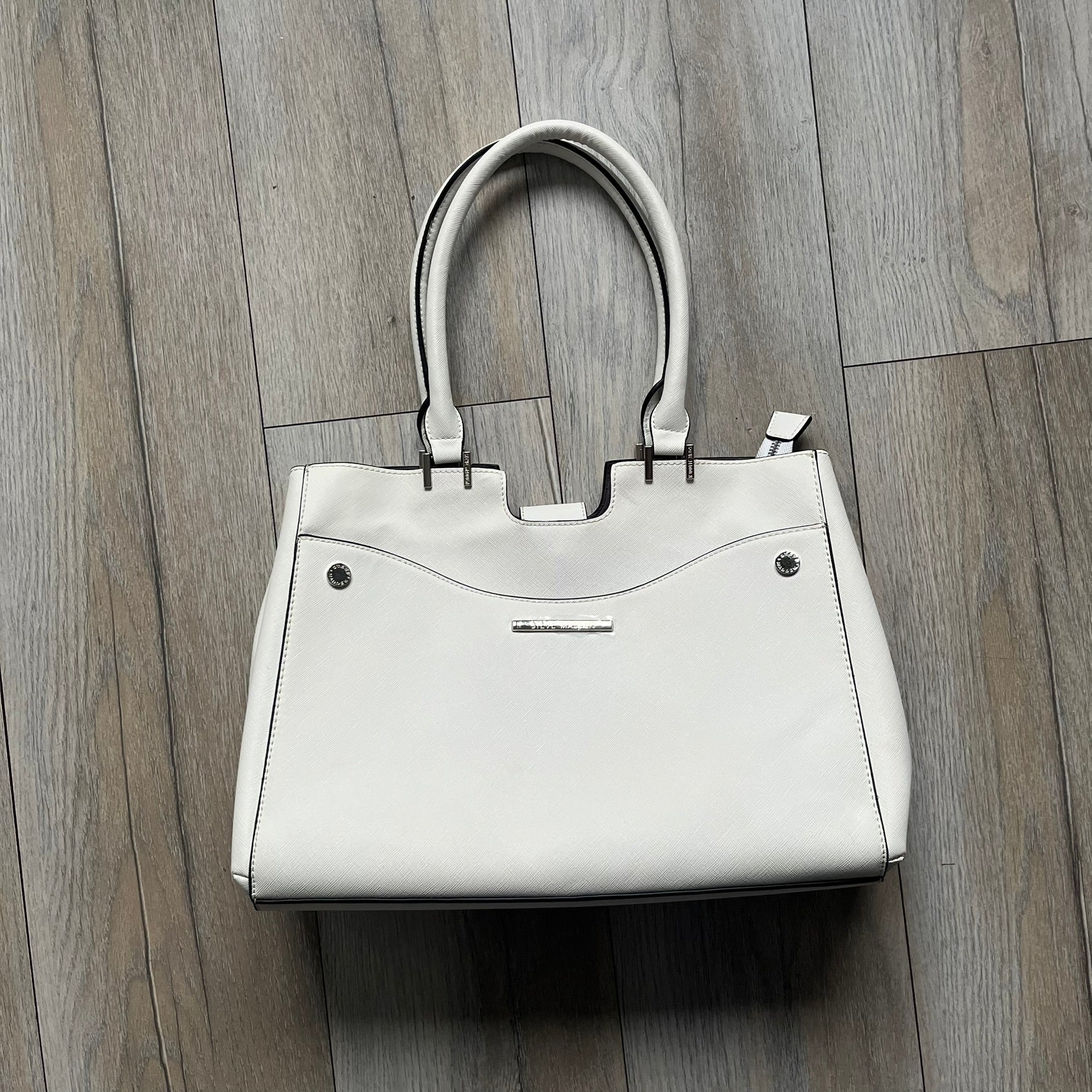 Steve Madden Removable Clutch White Handbag is a white leather tote featuring silver-tone hardware, including a front plaque and decorative buttons. The bag has two top handles and includes a removable inner clutch with silver Steve Madden branding. Designed for versatility, the clutch can be used separately or inside the main handbag.