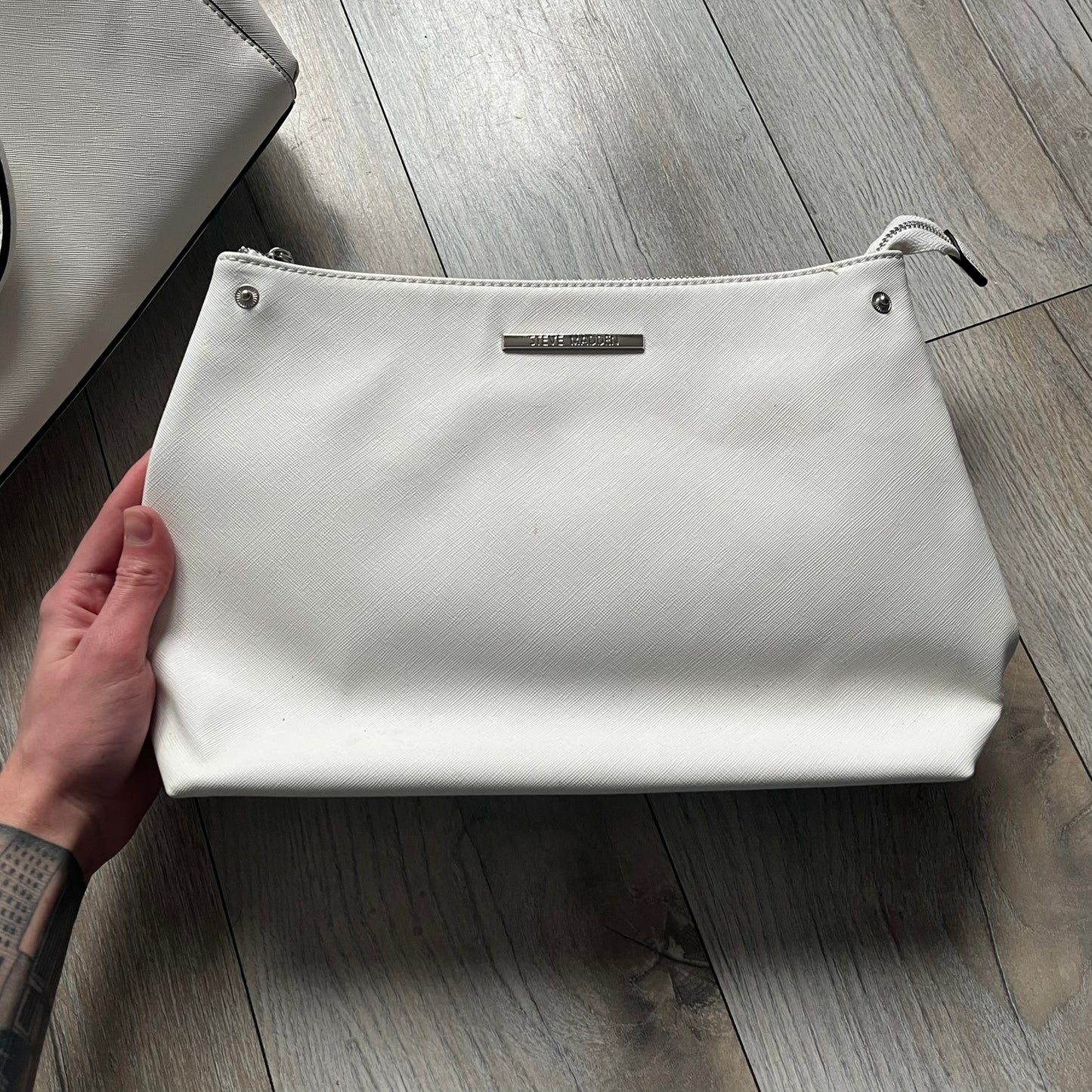 Steve Madden Removable Clutch White Handbag is a white leather tote featuring silver-tone hardware, including a front plaque and decorative buttons. The bag has two top handles and includes a removable inner clutch with silver Steve Madden branding. Designed for versatility, the clutch can be used separately or inside the main handbag.