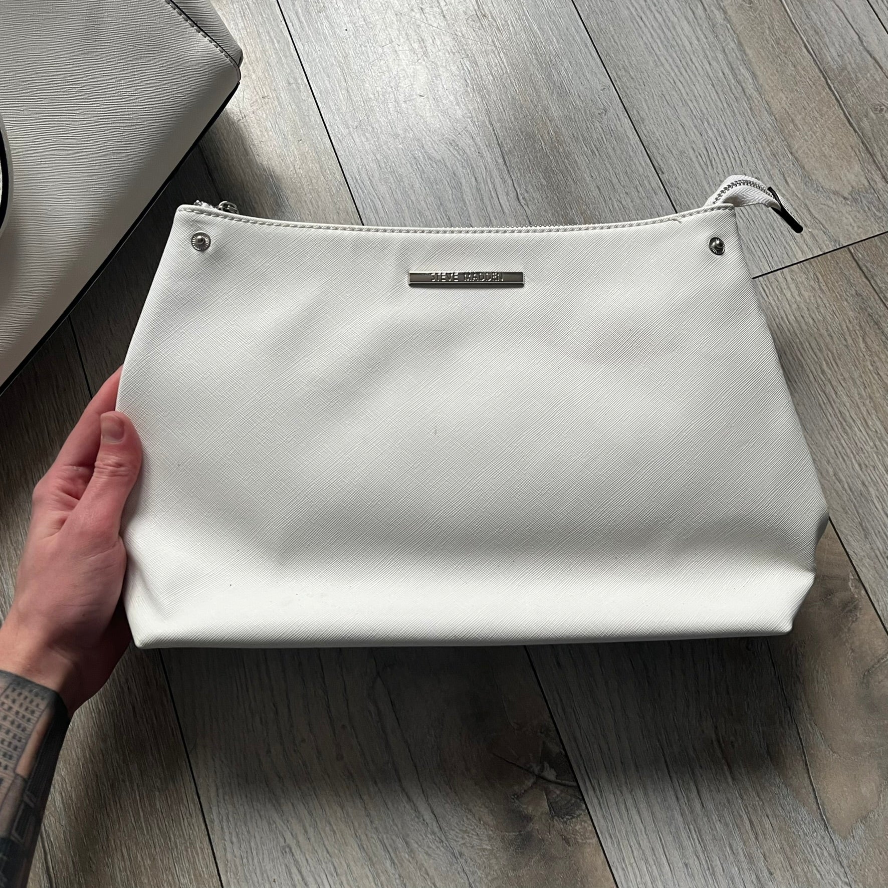 Steve Madden Removable Clutch White Handbag is a white leather tote featuring silver-tone hardware, including a front plaque and decorative buttons. The bag has two top handles and includes a removable inner clutch with silver Steve Madden branding. Designed for versatility, the clutch can be used separately or inside the main handbag.