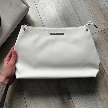 Steve Madden Removable Clutch White Handbag is a white leather tote featuring silver-tone hardware, including a front plaque and decorative buttons. The bag has two top handles and includes a removable inner clutch with silver Steve Madden branding. Designed for versatility, the clutch can be used separately or inside the main handbag.
