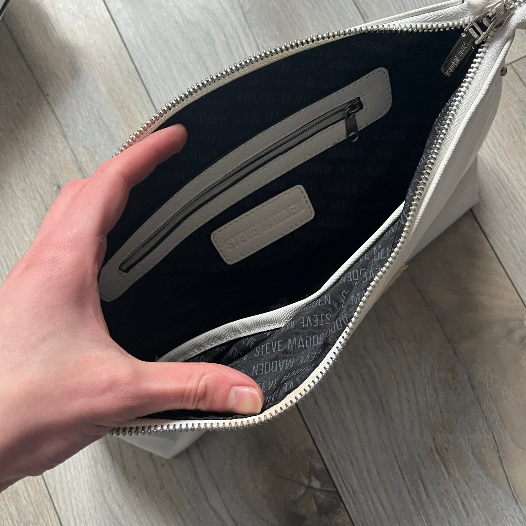 Steve Madden Removable Clutch White Handbag is a white leather tote featuring silver-tone hardware, including a front plaque and decorative buttons. The bag has two top handles and includes a removable inner clutch with silver Steve Madden branding. Designed for versatility, the clutch can be used separately or inside the main handbag.