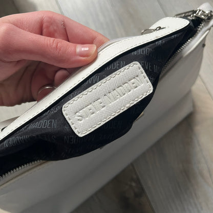 Steve Madden Removable Clutch White Handbag is a white leather tote featuring silver-tone hardware, including a front plaque and decorative buttons. The bag has two top handles and includes a removable inner clutch with silver Steve Madden branding. Designed for versatility, the clutch can be used separately or inside the main handbag.