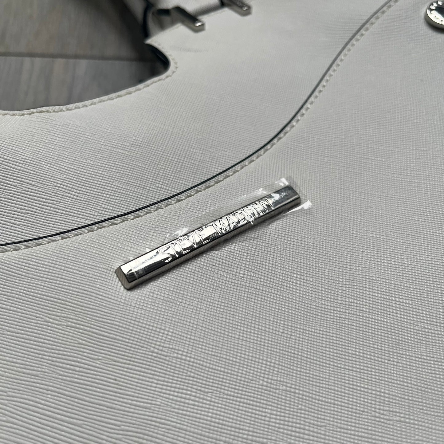 Steve Madden Removable Clutch White Handbag is a white leather tote featuring silver-tone hardware, including a front plaque and decorative buttons. The bag has two top handles and includes a removable inner clutch with silver Steve Madden branding. Designed for versatility, the clutch can be used separately or inside the main handbag.