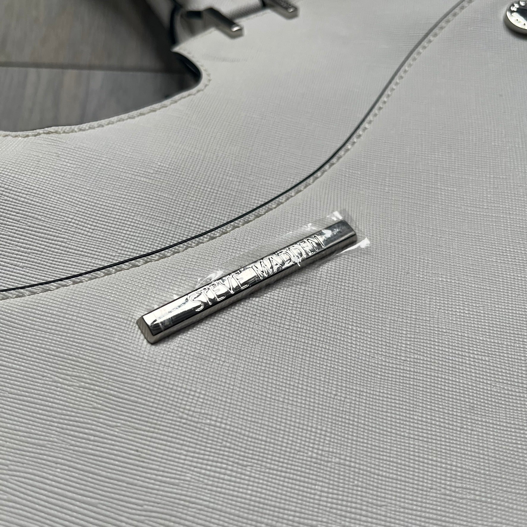 Steve Madden Removable Clutch White Handbag is a white leather tote featuring silver-tone hardware, including a front plaque and decorative buttons. The bag has two top handles and includes a removable inner clutch with silver Steve Madden branding. Designed for versatility, the clutch can be used separately or inside the main handbag.