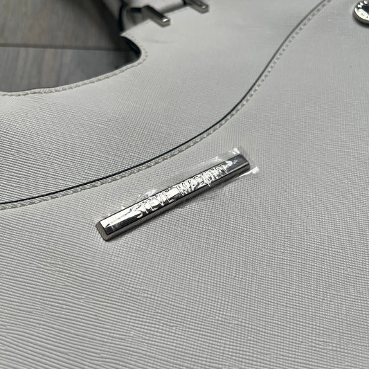 Steve Madden Removable Clutch White Handbag is a white leather tote featuring silver-tone hardware, including a front plaque and decorative buttons. The bag has two top handles and includes a removable inner clutch with silver Steve Madden branding. Designed for versatility, the clutch can be used separately or inside the main handbag.