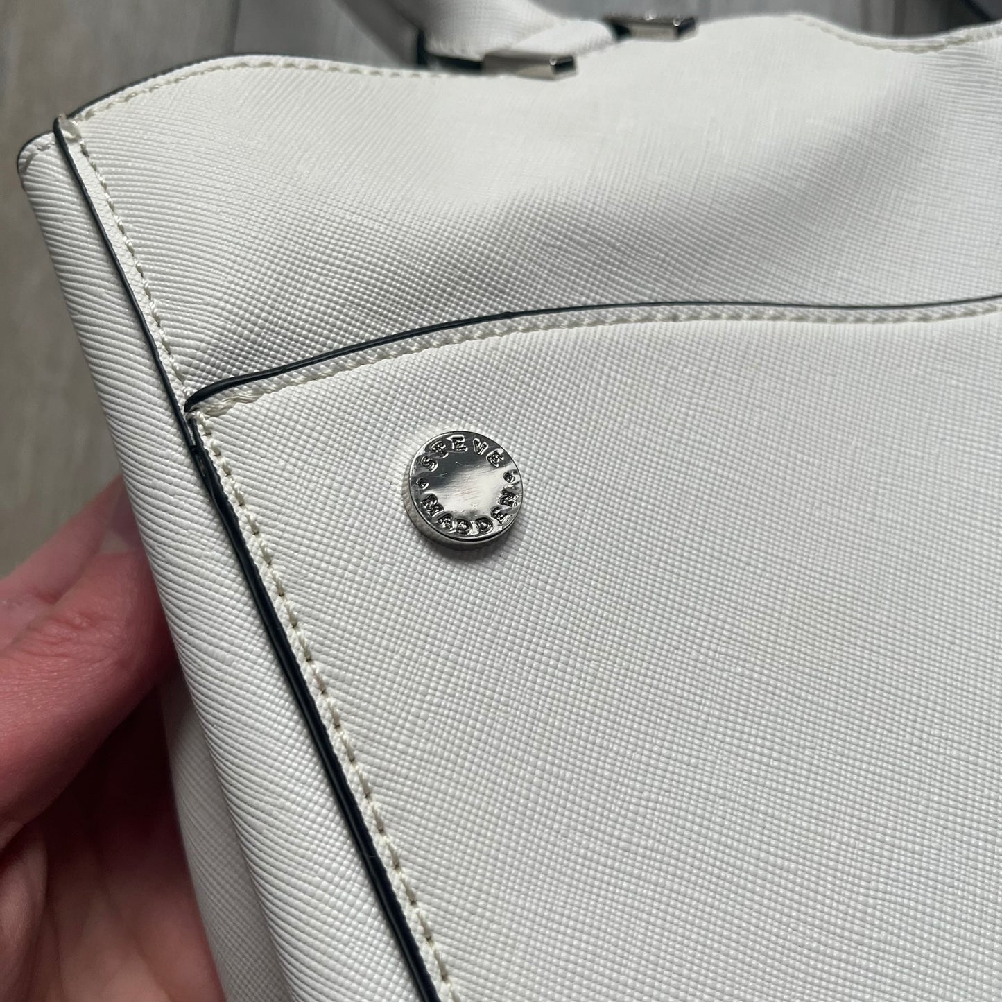 Steve Madden Removable Clutch White Handbag is a white leather tote featuring silver-tone hardware, including a front plaque and decorative buttons. The bag has two top handles and includes a removable inner clutch with silver Steve Madden branding. Designed for versatility, the clutch can be used separately or inside the main handbag.