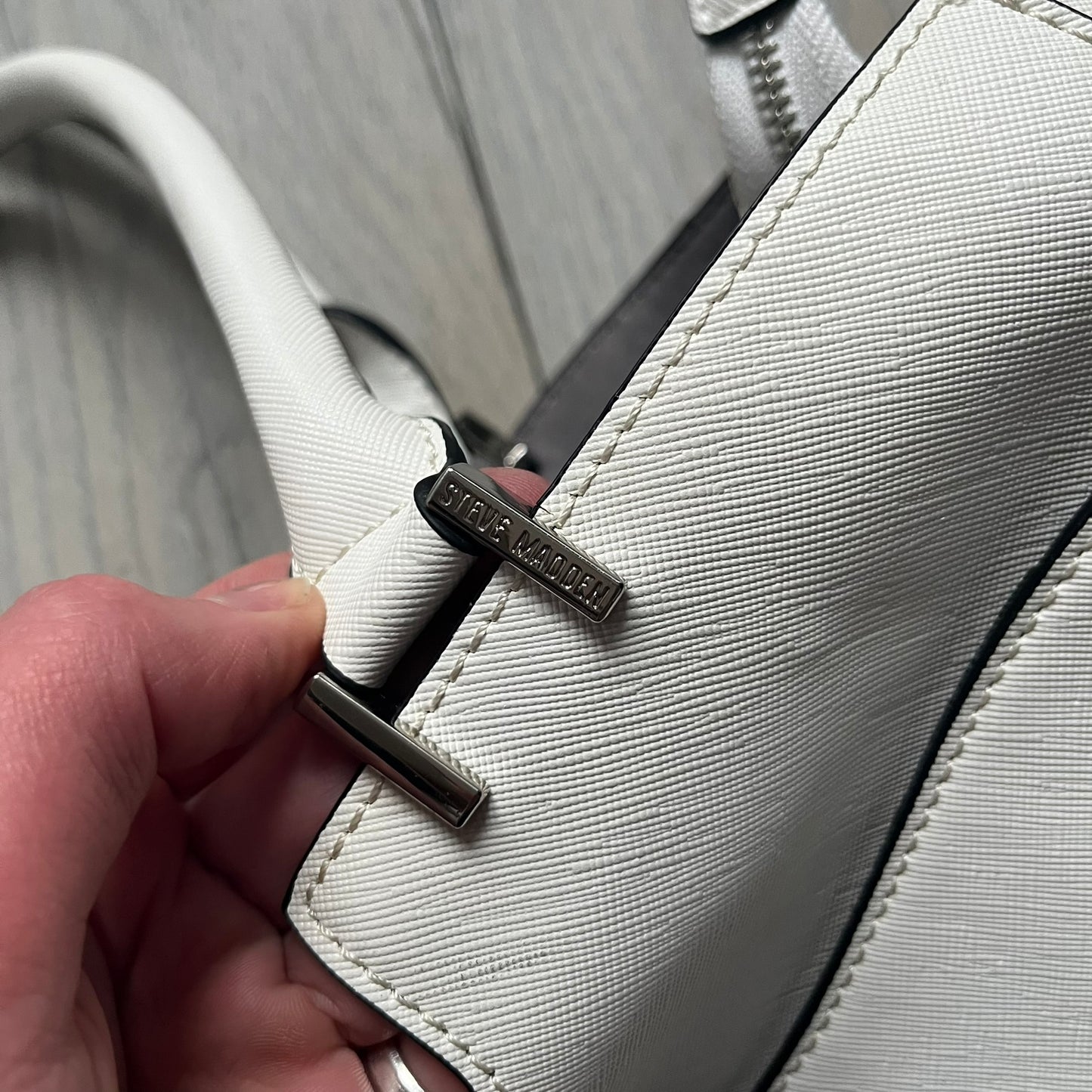 Steve Madden Removable Clutch White Handbag is a white leather tote featuring silver-tone hardware, including a front plaque and decorative buttons. The bag has two top handles and includes a removable inner clutch with silver Steve Madden branding. Designed for versatility, the clutch can be used separately or inside the main handbag.